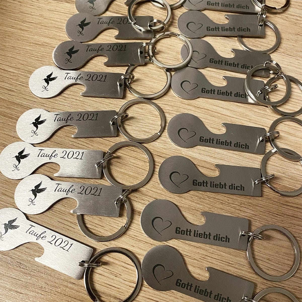 Mehrere Einkaufswagenlöser "Style&Open" Schlüsselanhänger mit "Taufe 2021" und "Gott liebt dich" eingraviert auf einer Holzoberfläche.