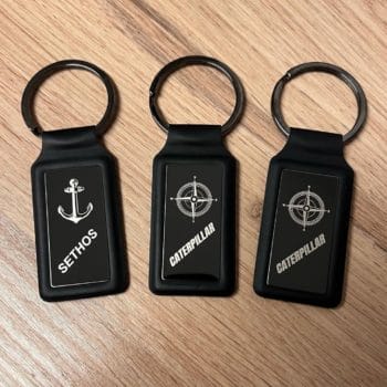 Drei schwarze "Dakar"-Schlüsselanhänger auf Holz, mit Caterpillar-Logos und einem Anker mit der Aufschrift "SETHOS".