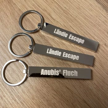 Drei "Blocky"-Schlüsselanhänger auf Holz: zwei mit der Gravur "Ländle Escape", einer mit "Anubis' Fluch".